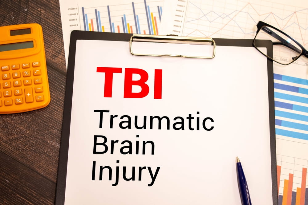 Clipboard labeled "TBI Traumatic Brain Injury" with data charts in the background