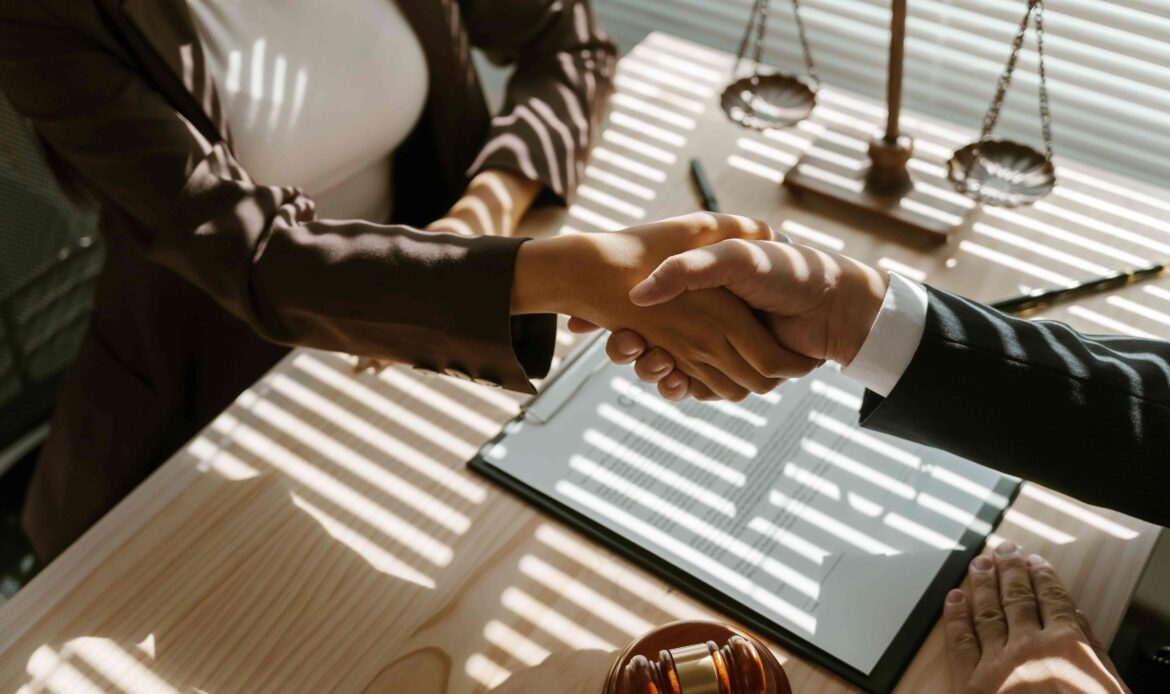 Lawyer and client shaking hands after successful legal agreement in office with gavel and scales of justice on desk.