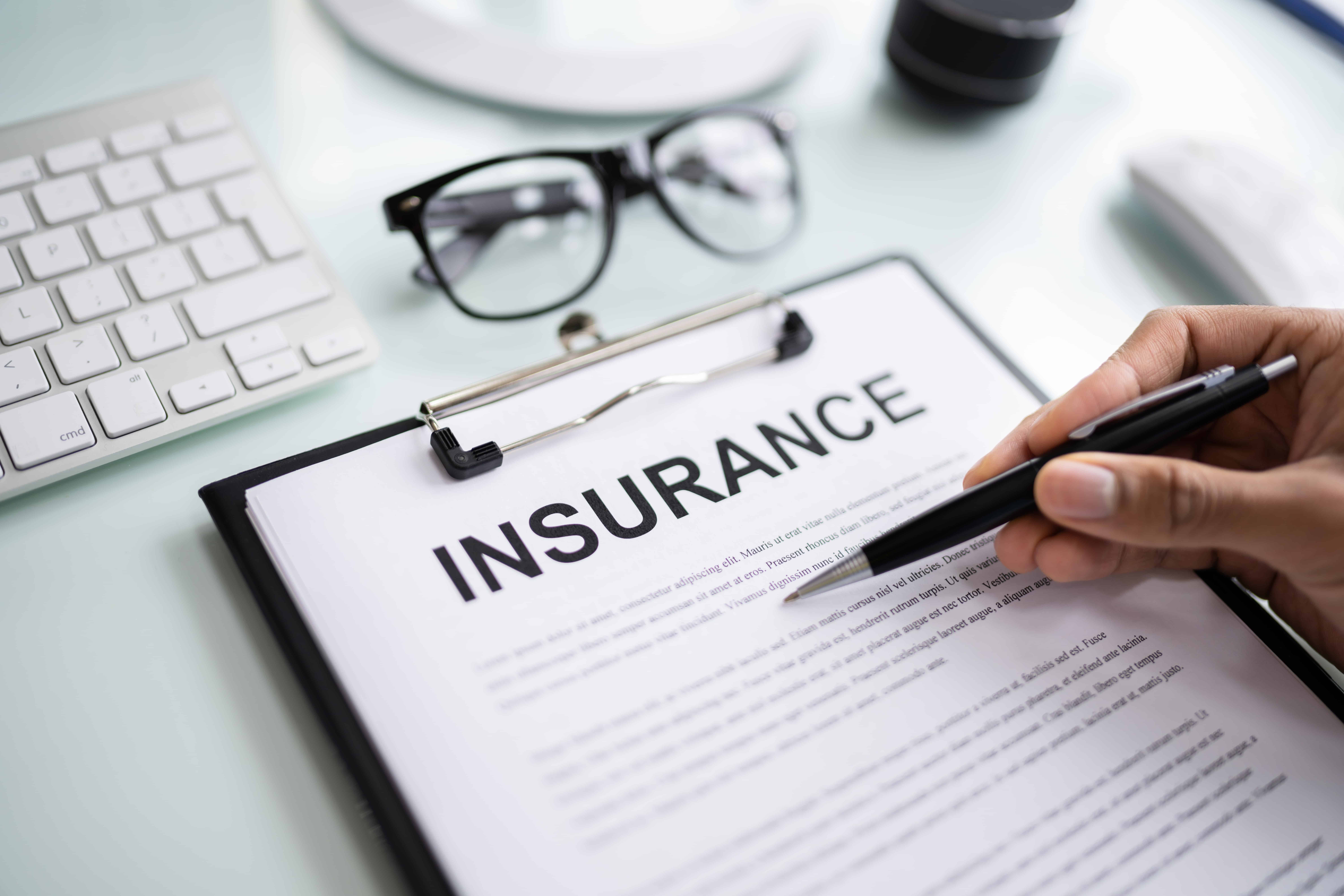 Close-up of person reviewing insurance policy document on clipboard with pen, glasses, and keyboard on desk. Close-up of person reviewing insurance policy document on clipboard with pen, glasses, and keyboard on desk.