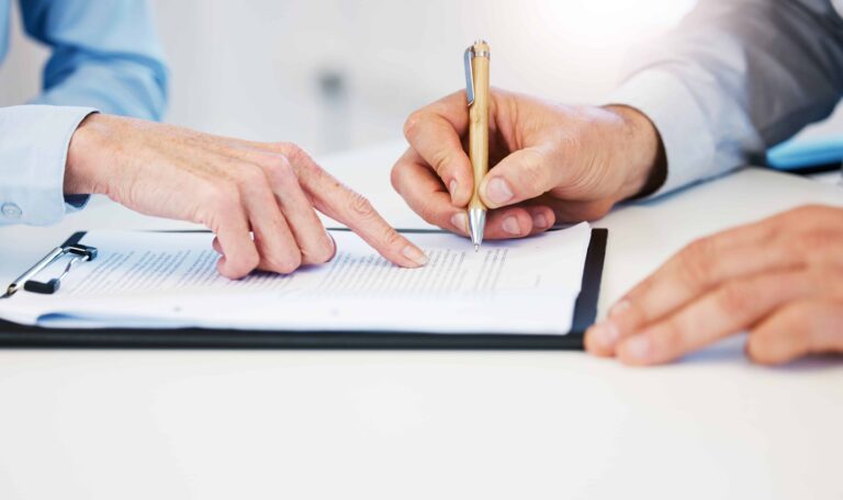 Person signing a legal document on a clipboard with guidance from another individual, representing contract review or agreement signing.