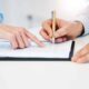 Person signing a legal document on a clipboard with guidance from another individual, representing contract review or agreement signing.