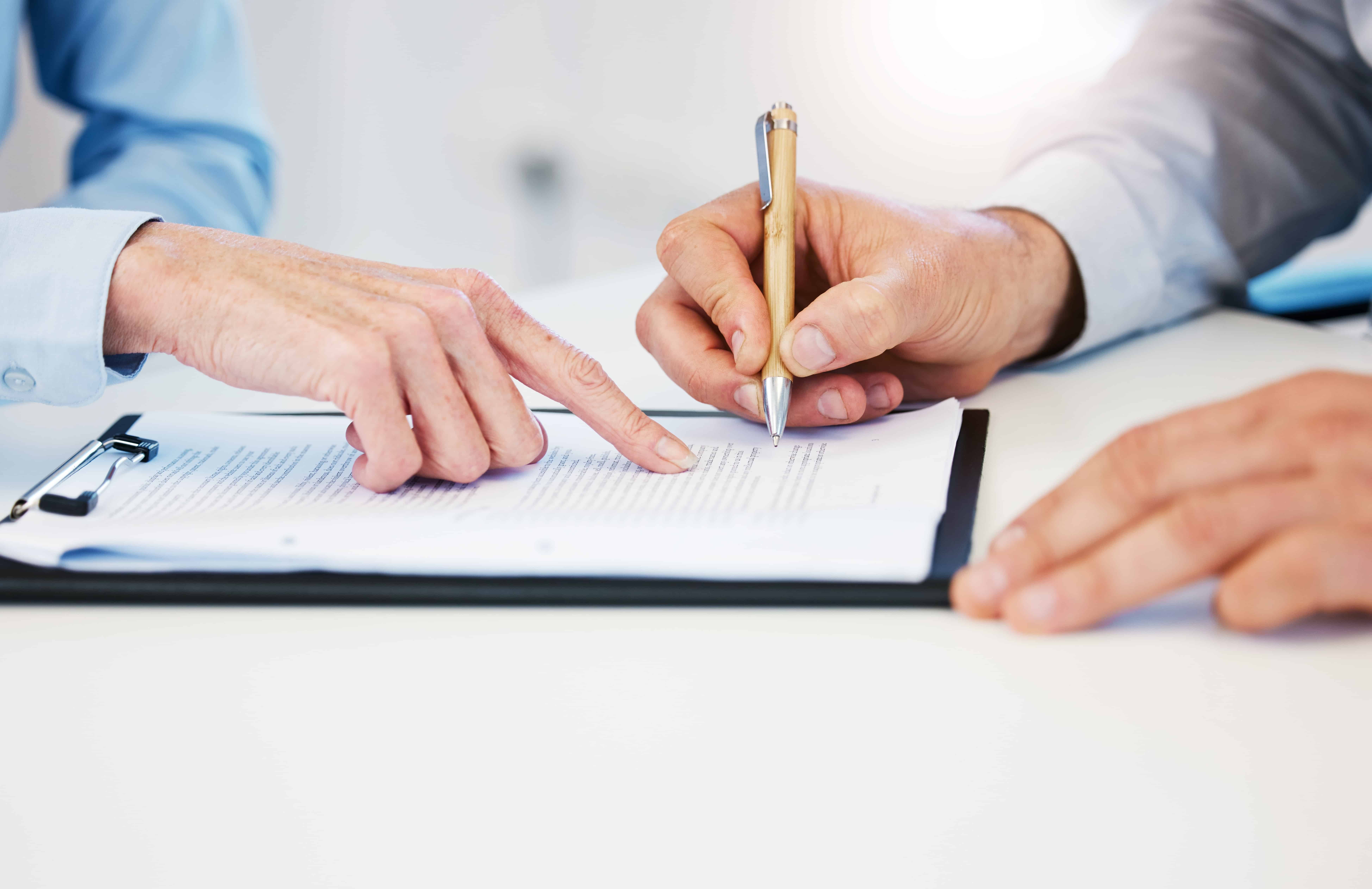 Person signing a legal document on a clipboard with guidance from another individual, representing contract review or agreement signing.