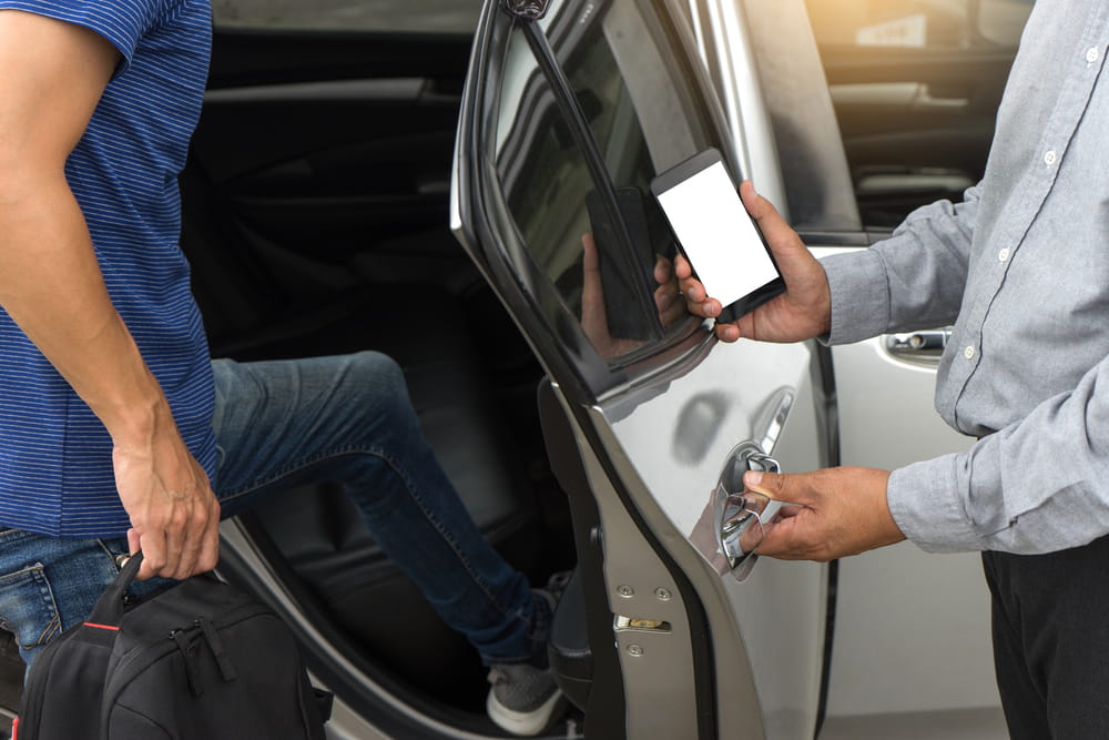 Passenger entering a rideshare vehicle with luggage while the driver holds a phone